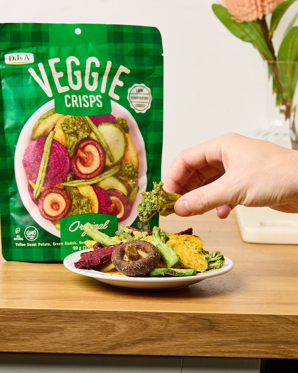 Hand picking a veggie crisp from a bowl next to a pack of DJ & A Veggie Crisps Original.
