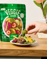 Hand picking a veggie crisp from a bowl next to a pack of DJ & A Veggie Crisps Original.